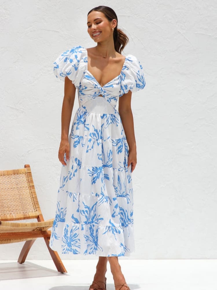 A woman wearing a white midi dress with a blue botanical print, featuring puff sleeves, a twist-front bodice with a cutout, and a tiered skirt, standing against a white textured wall.