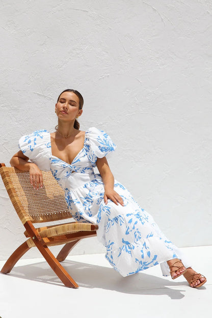 A woman wearing a white midi dress with a blue botanical print, featuring puff sleeves, a twist-front bodice with a cutout, and a tiered skirt, standing against a white textured wall.
