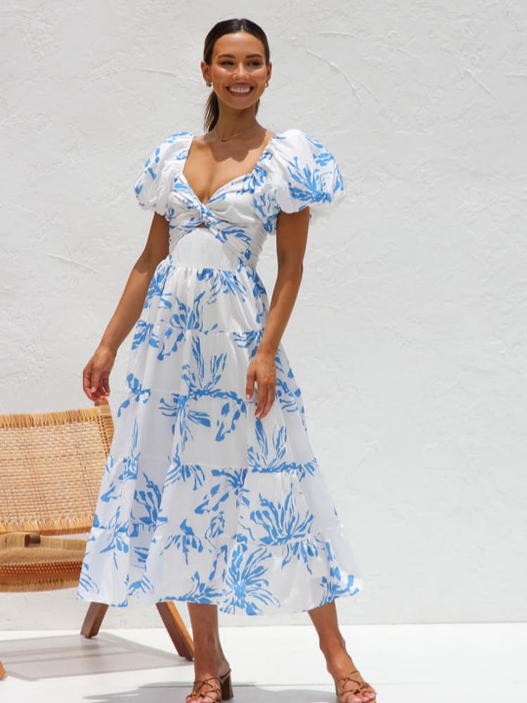 A woman wearing a white midi dress with a blue botanical print, featuring puff sleeves, a twist-front bodice with a cutout, and a tiered skirt, standing against a white textured wall.