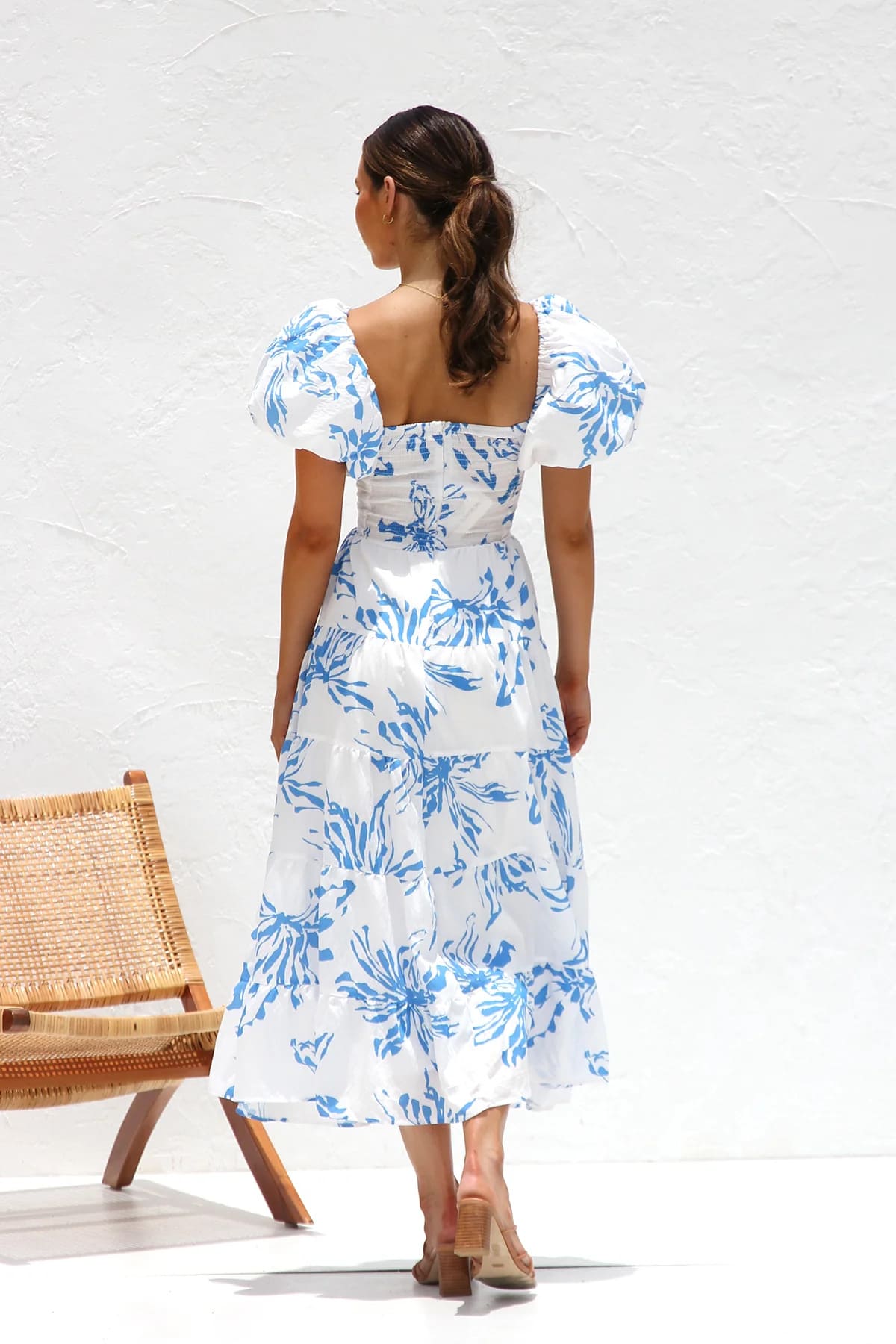 A woman wearing a white midi dress with a blue botanical print, featuring puff sleeves, a twist-front bodice with a cutout, and a tiered skirt, standing against a white textured wall.