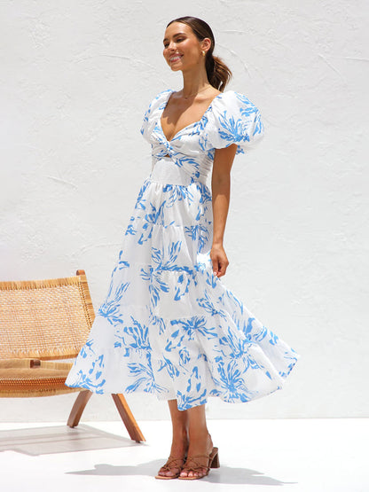 Blue - A woman wearing a white midi dress with a blue botanical print, featuring puff sleeves, a twist-front bodice with a cutout, and a tiered skirt, standing against a white textured wall.