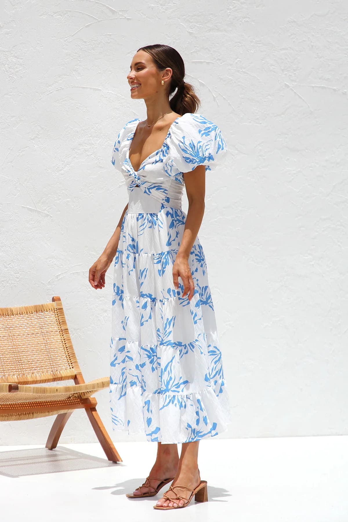 Blue - A woman wearing a white midi dress with a blue botanical print, featuring puff sleeves, a twist-front bodice with a cutout, and a tiered skirt, standing against a white textured wall.