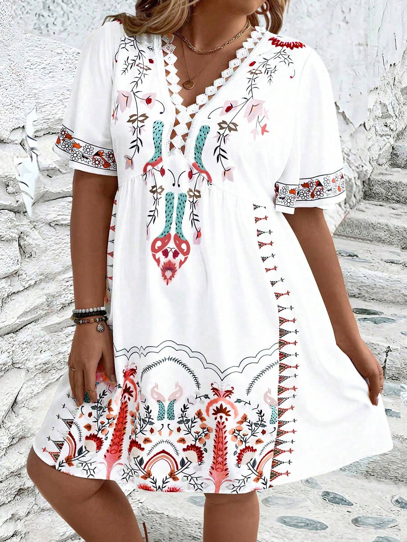 A woman wearing a white midi dress featuring a colorful folk-inspired floral print, a crochet-trimmed V-neckline, and short flutter sleeves, holding a woven straw clutch.
