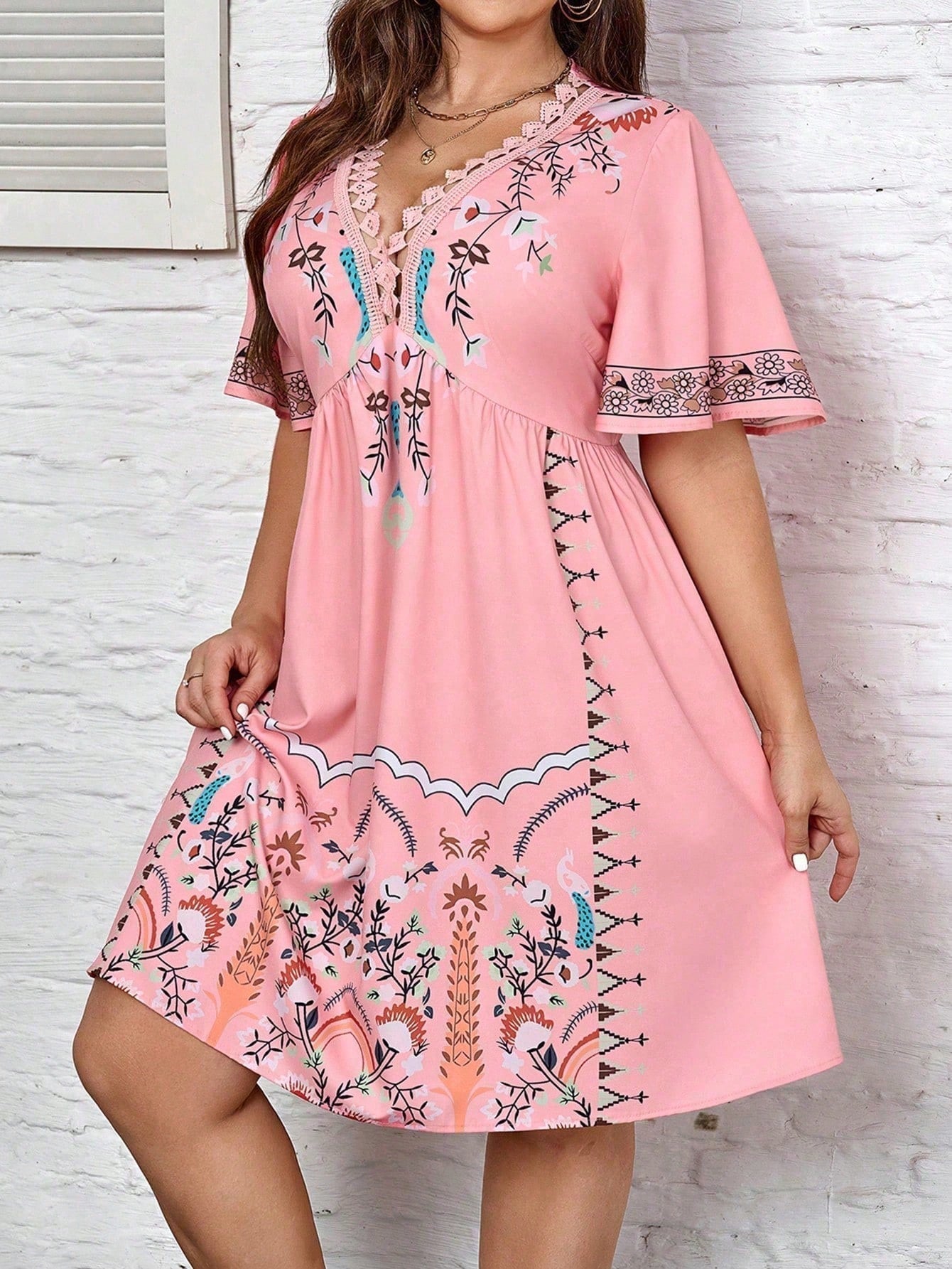White - A woman wearing a white midi dress featuring a colorful folk-inspired floral print, a crochet-trimmed V-neckline, and short flutter sleeves, holding a woven straw clutch.