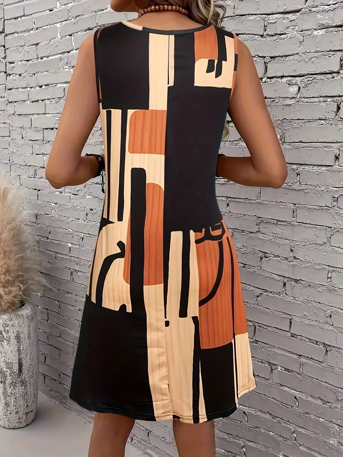 Chocolate - A woman wearing a sleeveless A-line shift dress featuring a bold black, white, and rust-orange geometric block print, styled with a bohemian wooden bead tassel necklace and a woven shoulder bag.