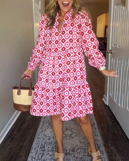 Pink - A woman wearing a pink and red geometric floral print cotton poplin tiered dress with long bishop sleeves and a ruffle neckline, paired with a straw handbag.