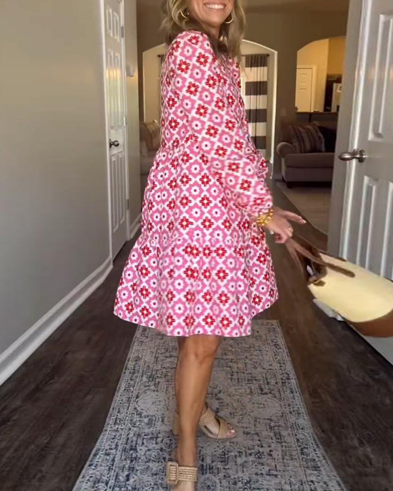 Pink - A woman wearing a pink and red geometric floral print cotton poplin tiered dress with long bishop sleeves and a ruffle neckline, paired with a straw handbag.