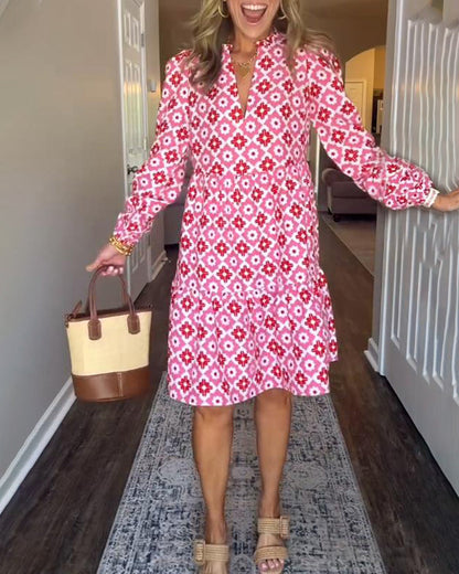 Pink - A woman wearing a pink and red geometric floral print cotton poplin tiered dress with long bishop sleeves and a ruffle neckline, paired with a straw handbag.