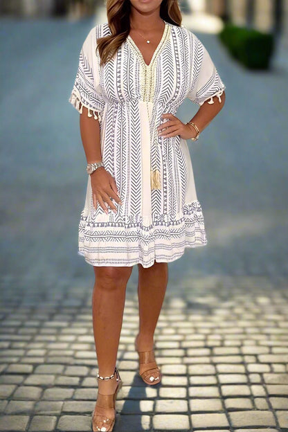 White/Blue - A woman wearing a white short-sleeved mini dress with a black geometric chevron print, featuring a V-neckline with embroidered trim, tassel-detailed flutter sleeves, and an adjustable drawstring waist.