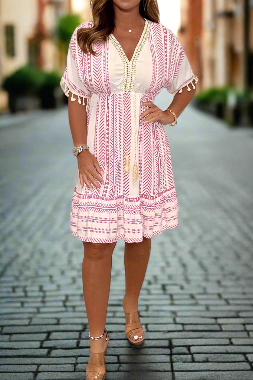 White/Pink - A woman wearing a white short-sleeved mini dress with a black geometric chevron print, featuring a V-neckline with embroidered trim, tassel-detailed flutter sleeves, and an adjustable drawstring waist.