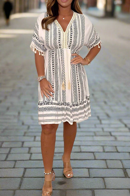White/Gray - A woman wearing a white short-sleeved mini dress with a black geometric chevron print, featuring a V-neckline with embroidered trim, tassel-detailed flutter sleeves, and an adjustable drawstring waist.