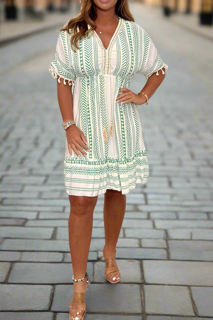 White/Green - A woman wearing a white short-sleeved mini dress with a black geometric chevron print, featuring a V-neckline with embroidered trim, tassel-detailed flutter sleeves, and an adjustable drawstring waist.