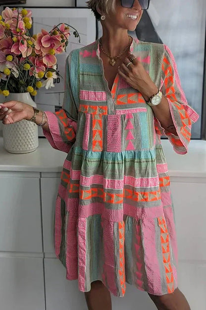 Colorful - A woman wearing a colorful geometric print tiered mini dress with a split neckline and three-quarter sleeves, featuring pink, orange, and green patterns on a textured fabric.
