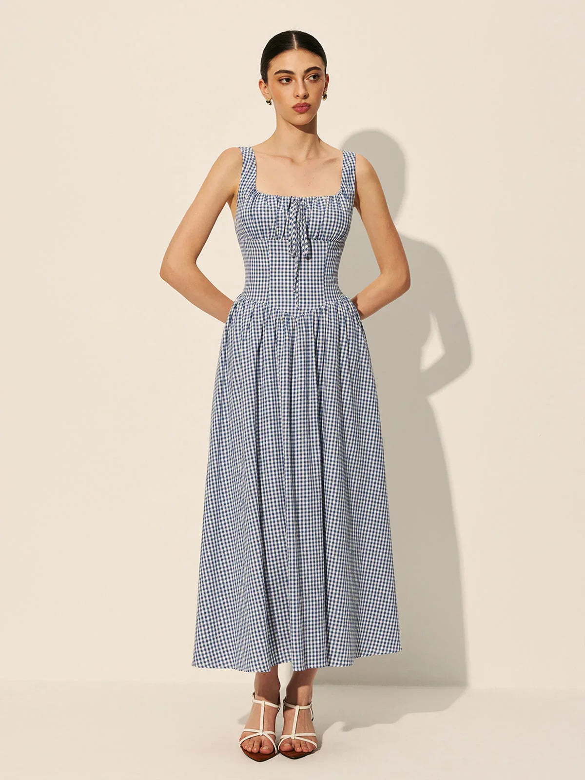 Blue - A woman wearing a blue and white gingham midi dress featuring a square neckline, corset-style bodice with a front tie, and a gathered A-line skirt.