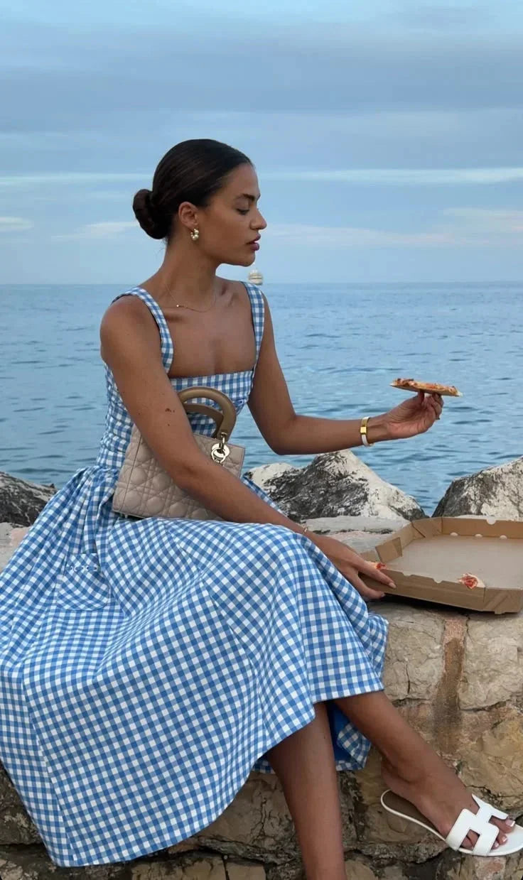 Hue - Woman wearing a blue and white gingham midi dress with a structured corset bodice and full pleated skirt, standing on a wooden dock.