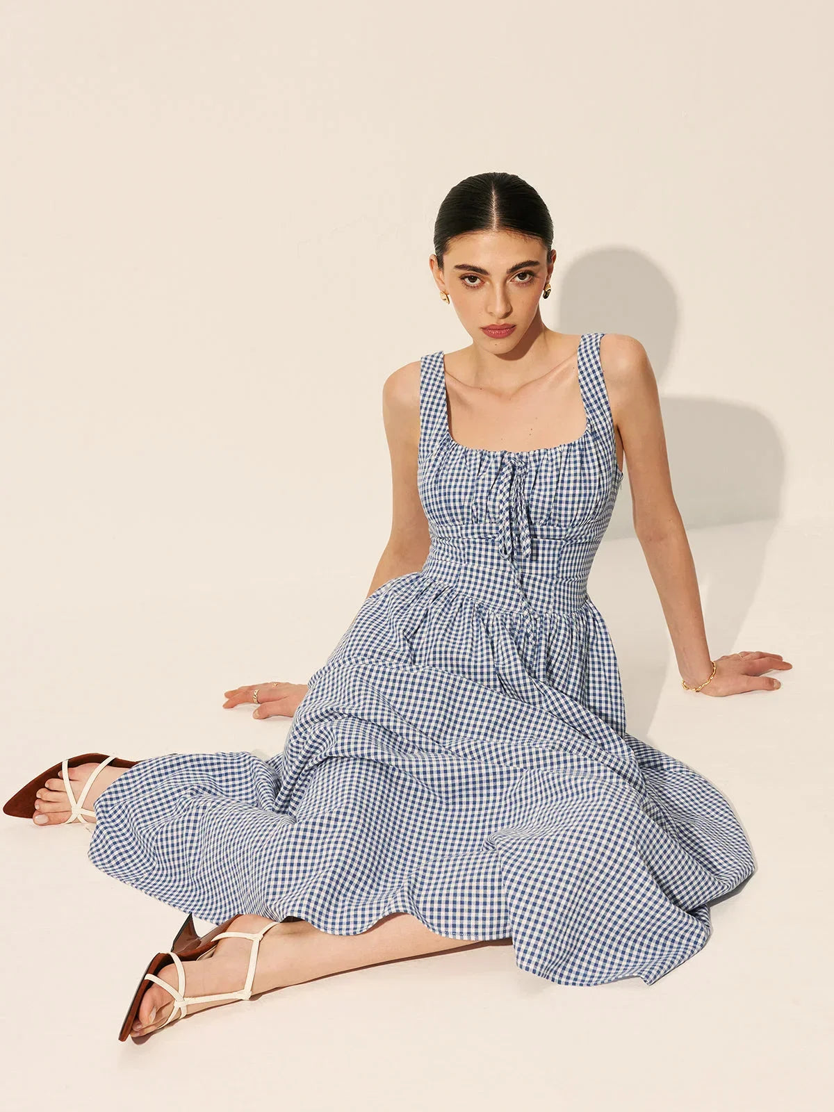 Blue - A woman wearing a blue and white gingham midi dress featuring a square neckline, corset-style bodice with a front tie, and a gathered A-line skirt.