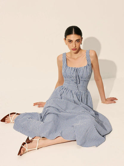 Blue - A woman wearing a blue and white gingham midi dress featuring a square neckline, corset-style bodice with a front tie, and a gathered A-line skirt.