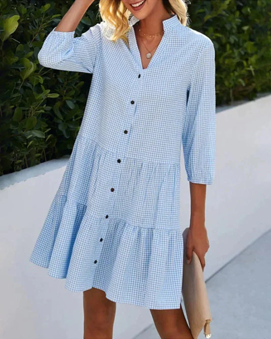 Light blue and white gingham mini dress featuring a split v-neckline, contrasting button-front closure, three-quarter sleeves, and a voluminous tiered skirt.