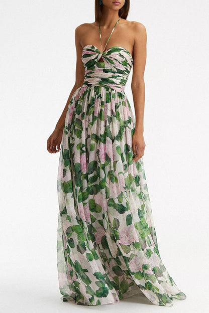 A woman wearing a floor-length chiffon maxi dress featuring a pink hydrangea and green leaf print, designed with a thin halter strap, a ruched sweetheart bodice, and a flowing skirt.