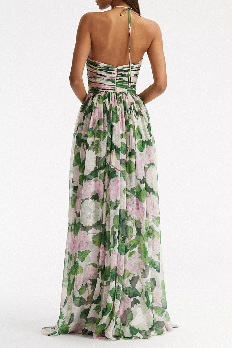 A woman wearing a floor-length chiffon maxi dress featuring a pink hydrangea and green leaf print, designed with a thin halter strap, a ruched sweetheart bodice, and a flowing skirt.