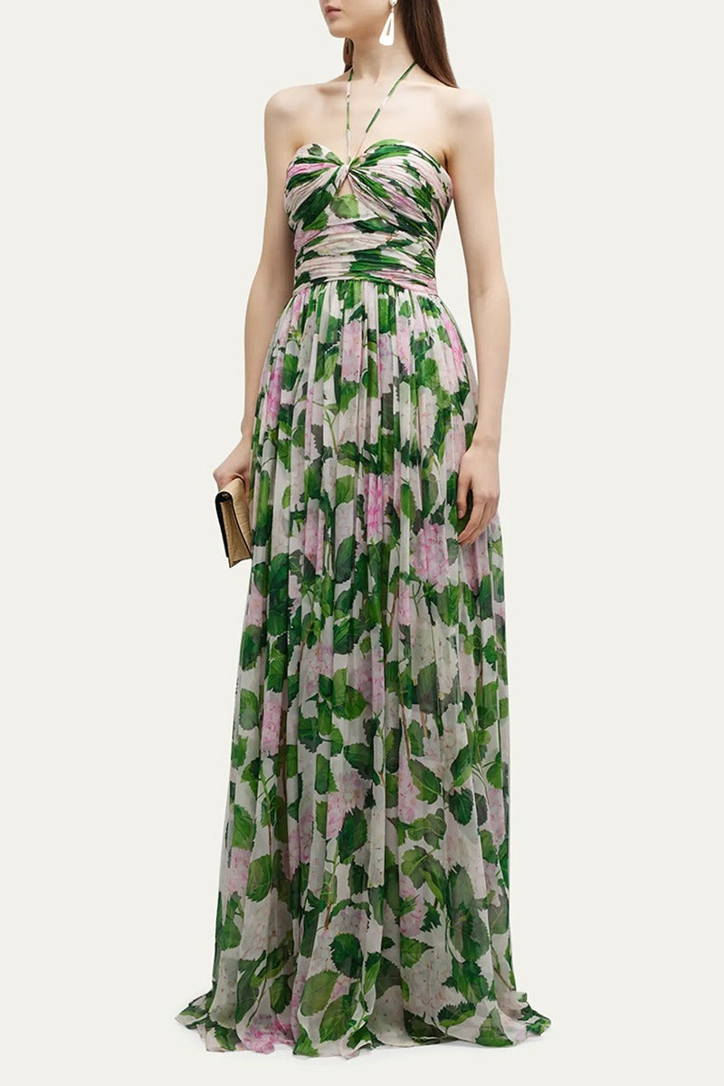 Green - A woman wearing a floor-length chiffon maxi dress featuring a pink hydrangea and green leaf print, designed with a thin halter strap, a ruched sweetheart bodice, and a flowing skirt.