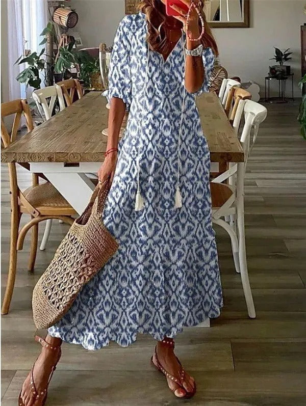 Blue - A woman wearing a blue and white ikat print maxi dress with a V-neckline, tassel ties, and short sleeves, holding a woven straw tote bag.