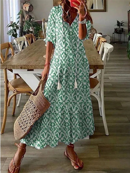 Green - A woman wearing a blue and white ikat print maxi dress with a V-neckline, tassel ties, and short sleeves, holding a woven straw tote bag.