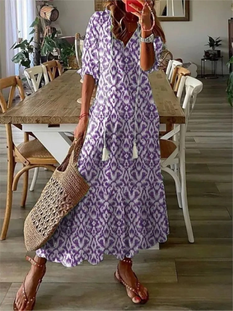 Purple - A woman wearing a blue and white ikat print maxi dress with a V-neckline, tassel ties, and short sleeves, holding a woven straw tote bag.