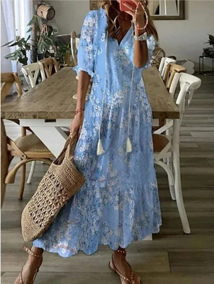 Light Blue - A woman wearing a blue and white ikat print maxi dress with a V-neckline, tassel ties, and short sleeves, holding a woven straw tote bag.