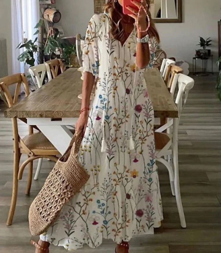 Floral - A woman wearing a blue and white ikat print maxi dress with a V-neckline, tassel ties, and short sleeves, holding a woven straw tote bag.