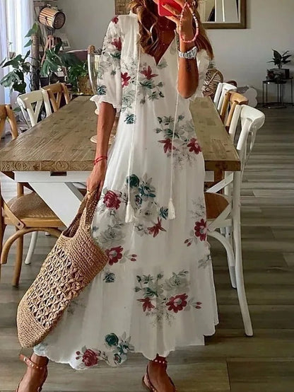 Flowers - A woman wearing a blue and white ikat print maxi dress with a V-neckline, tassel ties, and short sleeves, holding a woven straw tote bag.