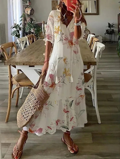 White - A woman wearing a blue and white ikat print maxi dress with a V-neckline, tassel ties, and short sleeves, holding a woven straw tote bag.