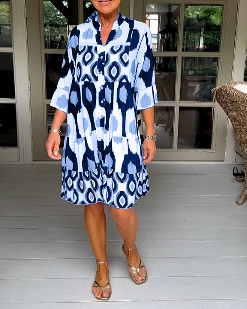 Blue - A woman wearing a knee-length, tiered tunic dress with a black, tan, and white geometric ikat print, featuring elbow-length sleeves with ruffled cuffs and a split V-neckline.