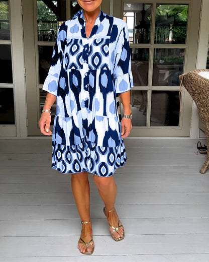 Blue - A woman wearing a knee-length, tiered tunic dress with a black, tan, and white geometric ikat print, featuring elbow-length sleeves with ruffled cuffs and a split V-neckline.