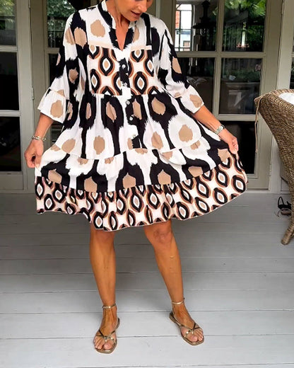White - A woman wearing a knee-length, tiered tunic dress with a black, tan, and white geometric ikat print, featuring elbow-length sleeves with ruffled cuffs and a split V-neckline.