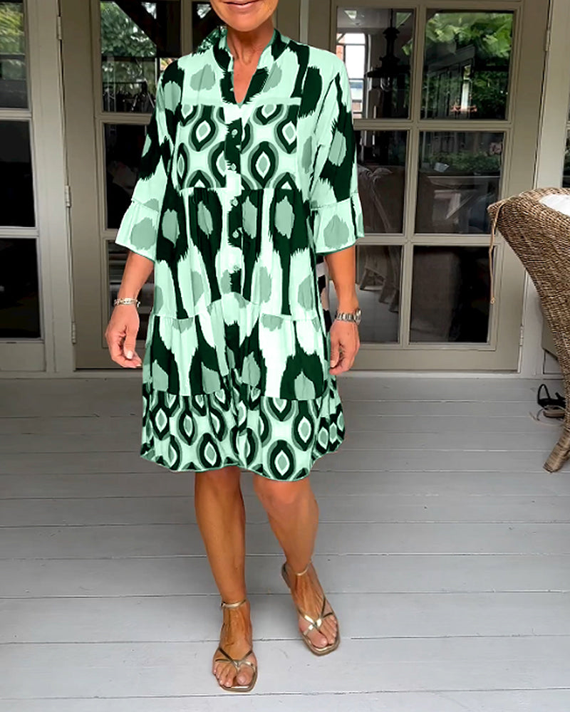 Green - A woman wearing a knee-length, tiered tunic dress with a black, tan, and white geometric ikat print, featuring elbow-length sleeves with ruffled cuffs and a split V-neckline.