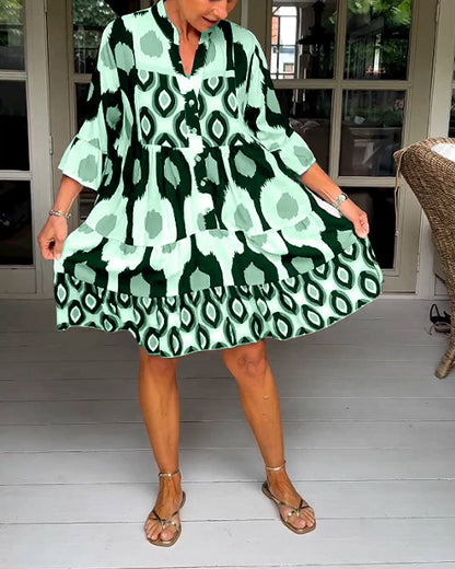 Green - A woman wearing a knee-length, tiered tunic dress with a black, tan, and white geometric ikat print, featuring elbow-length sleeves with ruffled cuffs and a split V-neckline.