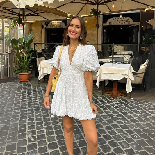 White - Woman wearing a white jacquard mini dress with puff sleeves and a bubble hem, standing on a cobblestone street in front of a restaurant.