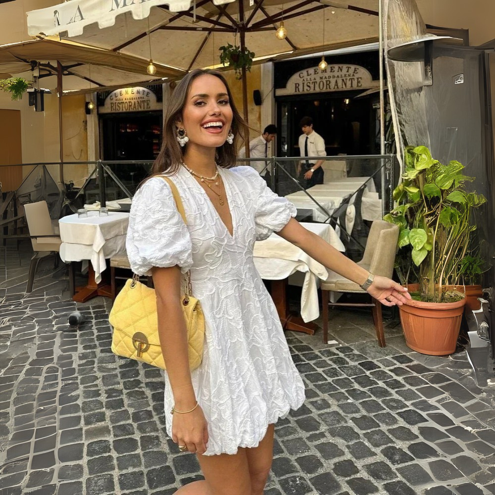 White - Woman wearing a white jacquard mini dress with puff sleeves and a bubble hem, standing on a cobblestone street in front of a restaurant.