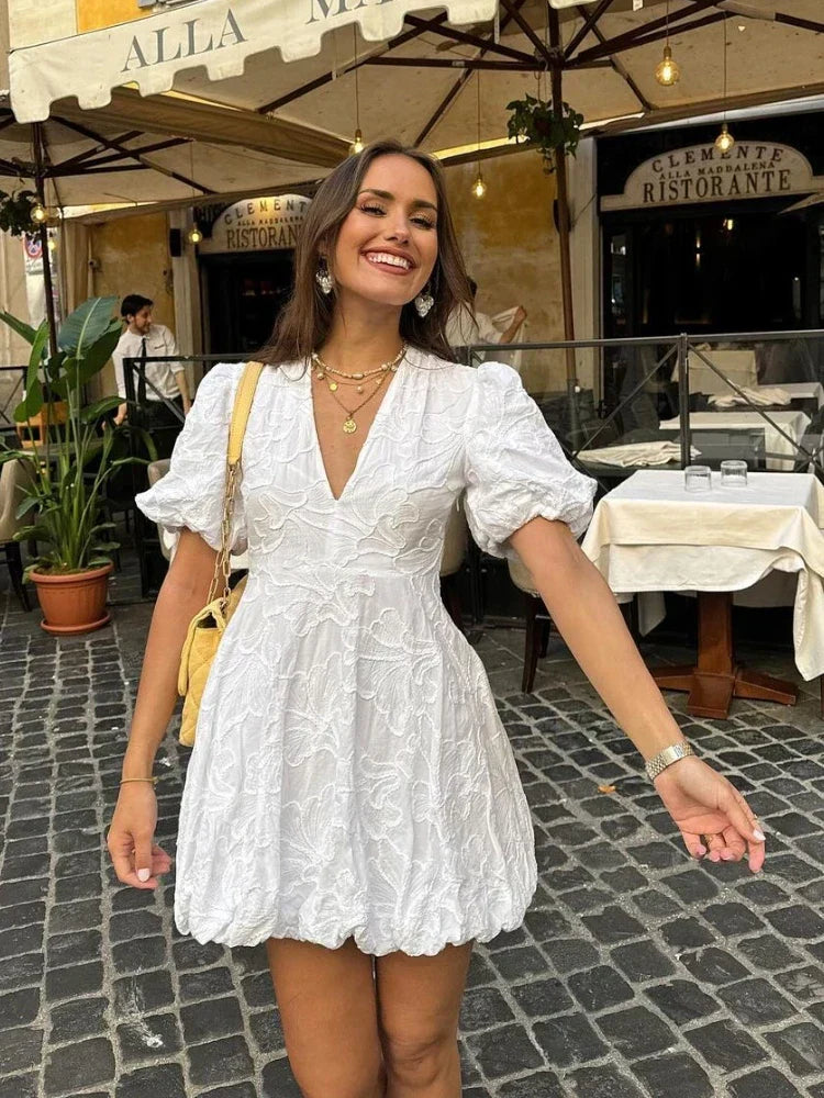 White - Woman wearing a white jacquard mini dress with puff sleeves and a bubble hem, standing on a cobblestone street in front of a restaurant.
