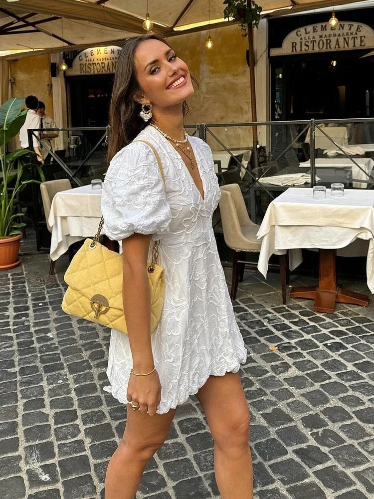 White - Woman wearing a white jacquard mini dress with puff sleeves and a bubble hem, standing on a cobblestone street in front of a restaurant.