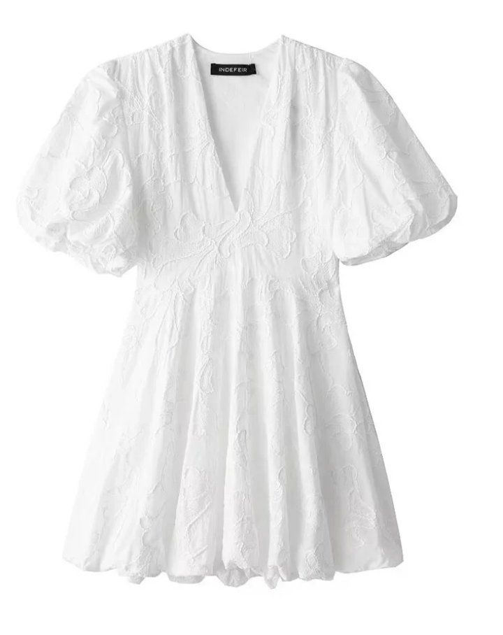 White - Woman wearing a white jacquard mini dress with puff sleeves and a bubble hem, standing on a cobblestone street in front of a restaurant.