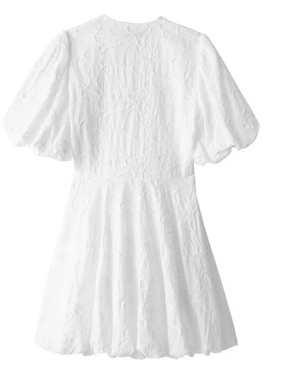 White - Woman wearing a white jacquard mini dress with puff sleeves and a bubble hem, standing on a cobblestone street in front of a restaurant.