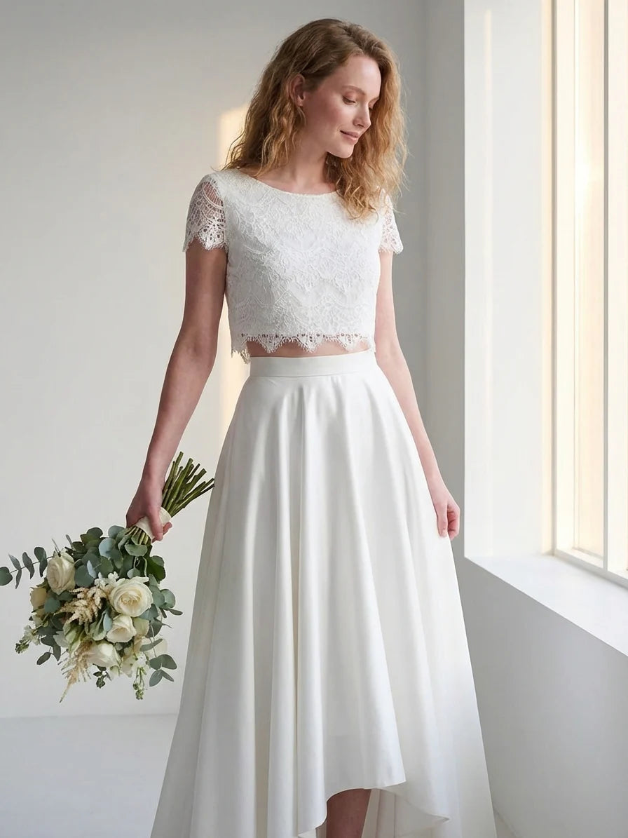 White - A woman wearing an elegant two-piece white bridal set featuring a short-sleeve cropped lace top and a high-waisted high-low maxi skirt while holding a bouquet of white roses.