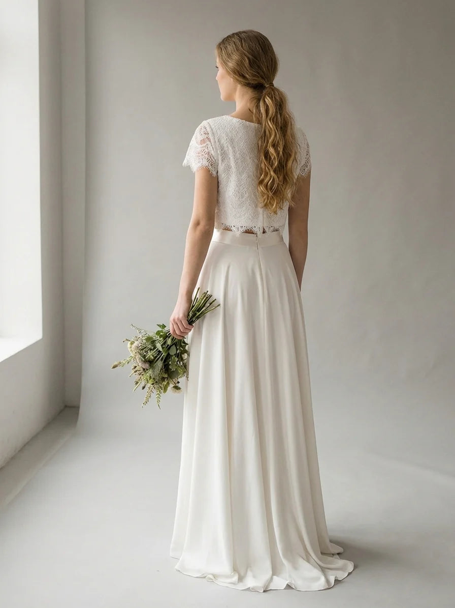 White - A woman wearing an elegant two-piece white bridal set featuring a short-sleeve cropped lace top and a high-waisted high-low maxi skirt while holding a bouquet of white roses.