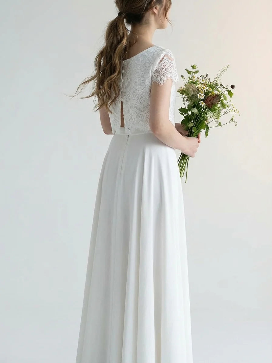 White - A woman wearing an elegant two-piece white bridal set featuring a short-sleeve cropped lace top and a high-waisted high-low maxi skirt while holding a bouquet of white roses.