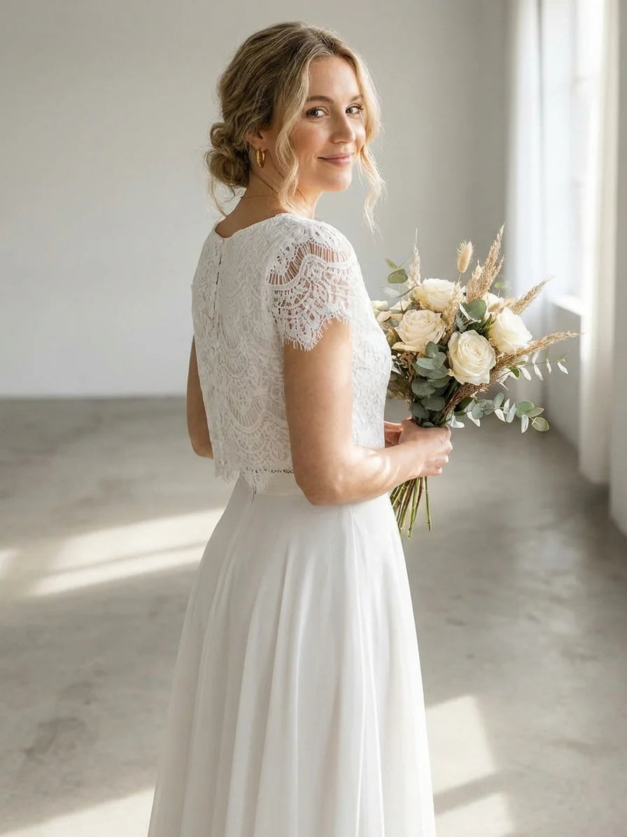 White - A woman wearing an elegant two-piece white bridal set featuring a short-sleeve cropped lace top and a high-waisted high-low maxi skirt while holding a bouquet of white roses.