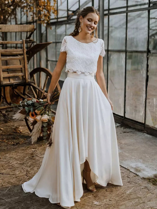 White - Woman wearing a white two-piece maxi dress featuring a short-sleeve floral lace crop top with a scalloped hem and a high-waisted, flowing skirt with a high-low hemline.