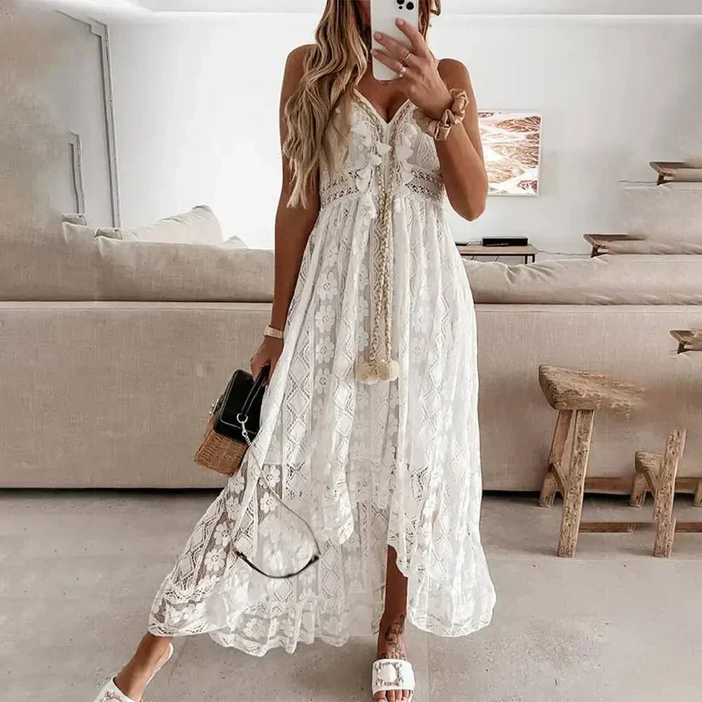 White - A woman wearing a white sleeveless high-low maxi dress made of paneled floral lace, featuring a V-neckline, braided tassel ties at the waist, and a flowing hemline.