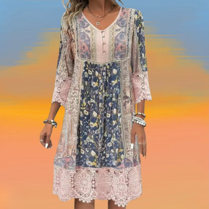 A woman wearing a bohemian-style mixed floral and paisley print shift dress with pink crochet lace trim on the 3/4 sleeves and hem, featuring a V-neckline with buttons, set against a scenic green hill and lake background.